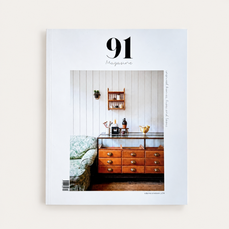 A magazine titled '91 Magazine' shown upright against a plain light background. The cover features a photograph of a cozy interior scene with a wooden chest of drawers, a green patterned armchair, and a white paneled wall.