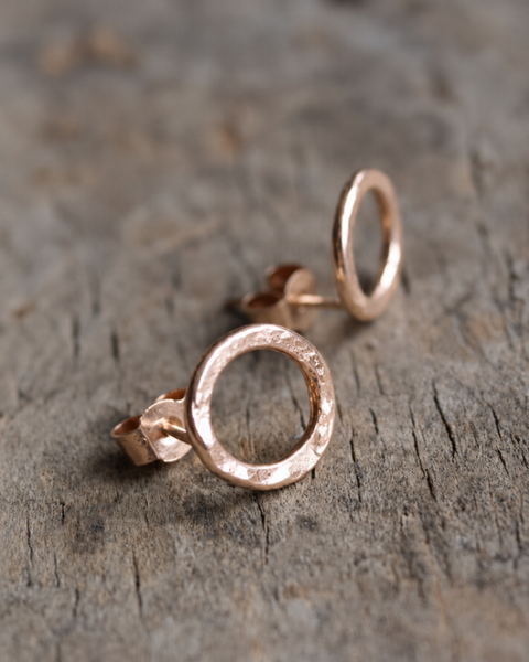 Close-up of handmade rose gold vermeil hammered circle stud earrings by Oriana Evans, showing the textured finish, posts and butterfly backs on a weathered wooden surface.