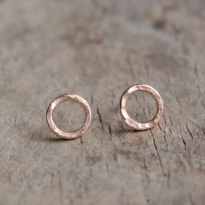 Pair of handmade rose gold vermeil circle stud earrings by Oriana Evans, featuring a delicate hammered finish, photographed on a natural wooden surface.