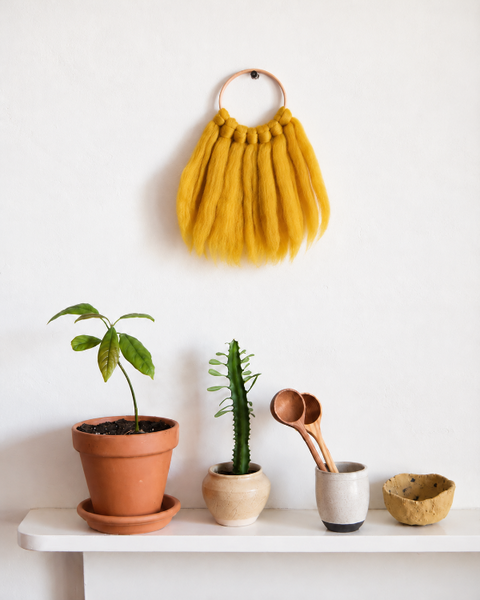 Mini mustard yellow merino wool wall hanging displayed above a wooden shelf with potted plants and decorative objects.