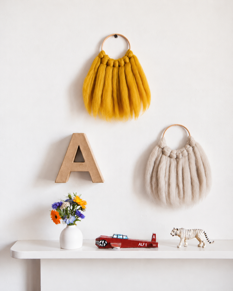 Handmade mustard yellow wool wall hanging displayed in a nursery alongside a natural wool wall hanging, wooden letter, flowers and small toys.