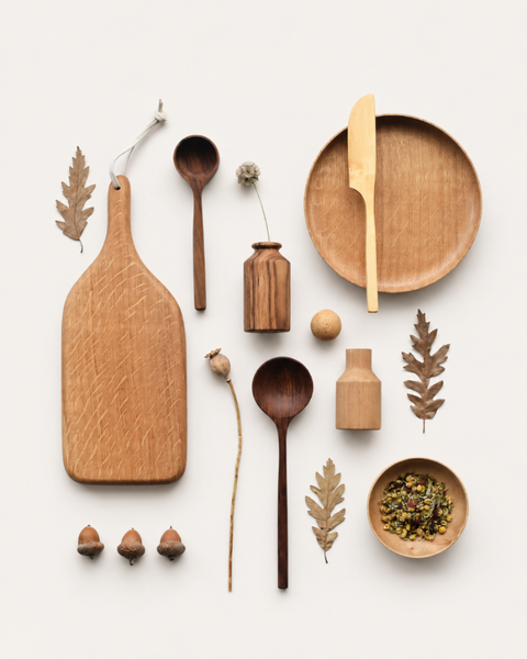 A styled flat-lay of natural wood kitchen and dining accessories on a light off-white background, including a large cutting board, two wooden spoons, a round plate with a knife, small wooden bottles, acorns, leaves, and a small bowl of seeds.