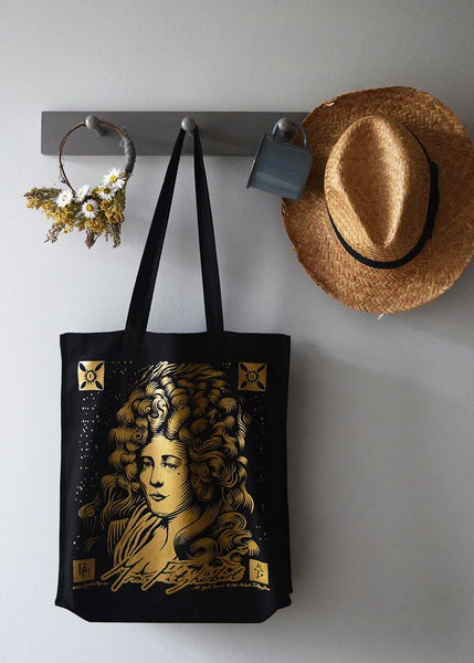 Black Mrs Fitzherbert canvas tote bag with gold illustration by Malcolm Trollope-Davis, hanging on wall hooks beside a straw hat, ceramic mug and dried flowers.